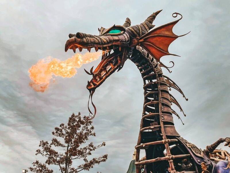 Une grande sculpture de dragon mécanique aux détails complexes, exhalant du feu sur un ciel nuageux, située dans un environnement extérieur entouré d'arbres à Disneyland Paris.