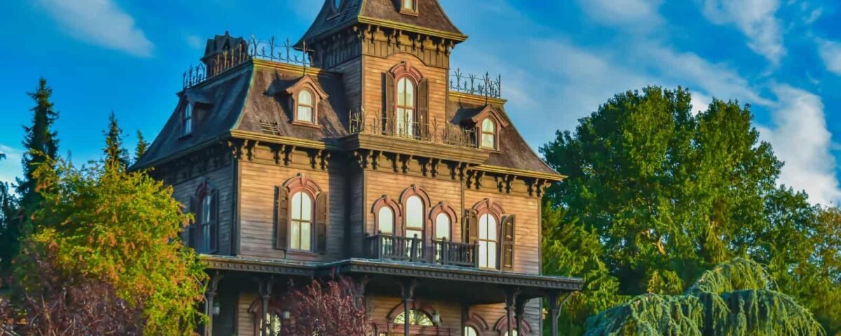 Un grand et étrange manoir victorien peint dans des tons sombres avec des boiseries complexes et entouré d'une verdure luxuriante et envahie par la végétation sous un ciel bleu vif avec des nuages duveteux, rappelant une attraction de Disneyland Paris.