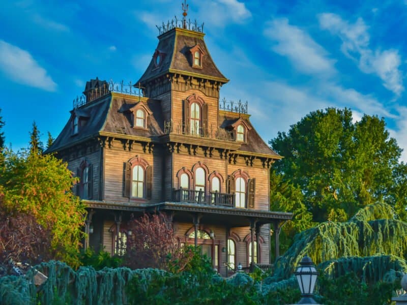 Un grand et étrange manoir victorien peint dans des tons sombres avec des boiseries complexes et entouré d'une verdure luxuriante et envahie par la végétation sous un ciel bleu vif avec des nuages duveteux, rappelant une attraction de Disneyland Paris.