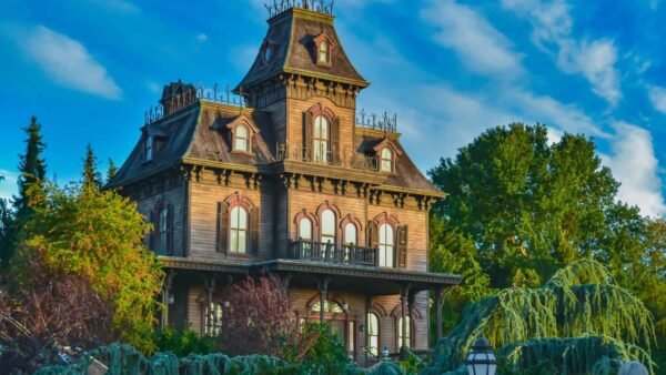 Un grand et étrange manoir victorien peint dans des tons sombres avec des boiseries complexes et entouré d'une verdure luxuriante et envahie par la végétation sous un ciel bleu vif avec des nuages duveteux, rappelant une attraction de Disneyland Paris.
