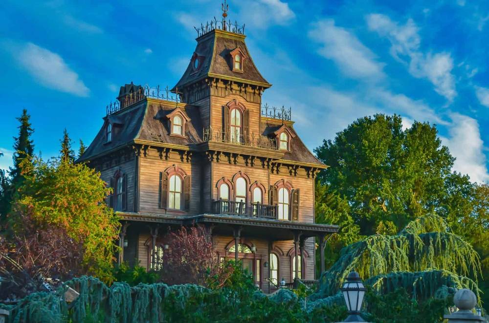 Un grand et étrange manoir victorien peint dans des tons sombres avec des boiseries complexes et entouré d'une verdure luxuriante et envahie par la végétation sous un ciel bleu vif avec des nuages duveteux, rappelant une attraction de Disneyland Paris.