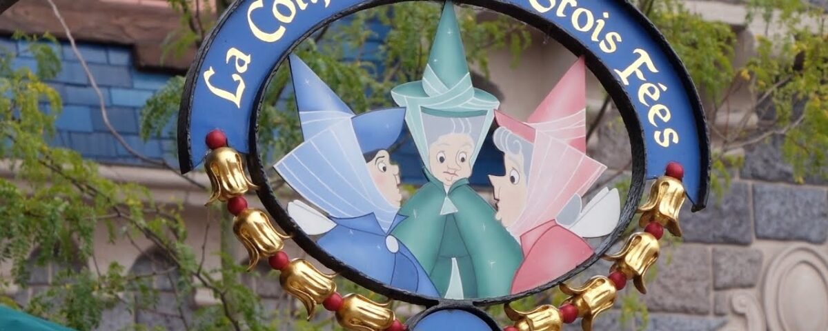 Une pancarte suspendue représentant l'illustration de trois fées marraines de la Belle au bois dormant sous le titre français "La Confiserie des 3 Fées", entourée d'un cadre décoratif doré, avec un Kinder