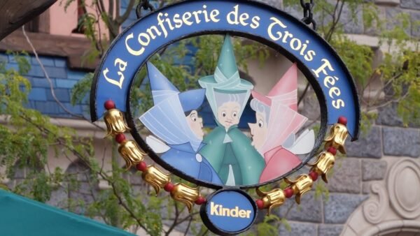 Une pancarte suspendue représentant l'illustration de trois fées marraines de la Belle au bois dormant sous le titre français "La Confiserie des 3 Fées", entourée d'un cadre décoratif doré, avec un Kinder