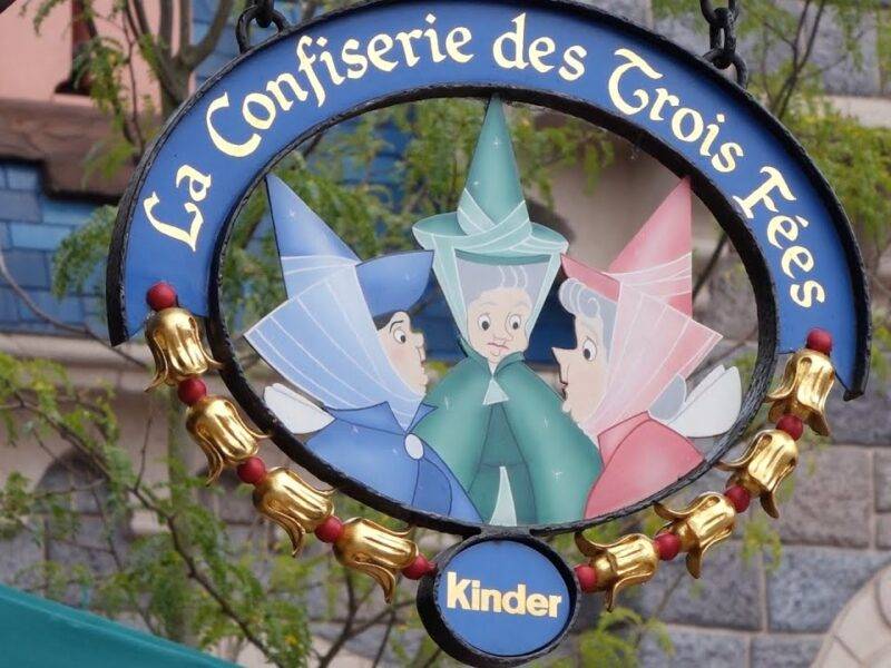 Une pancarte suspendue représentant l'illustration de trois fées marraines de la Belle au bois dormant sous le titre français "La Confiserie des 3 Fées", entourée d'un cadre décoratif doré, avec un Kinder