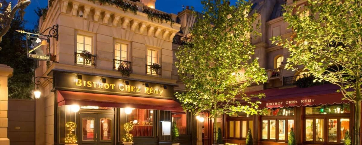 Une charmante vue nocturne du "Bistrot Chez Rémy", l'une des principales attractions de Disneyland Paris, avec des intérieurs chaleureusement éclairés et des lampadaires ornés, entouré d'arbres verts luxuriants dans un