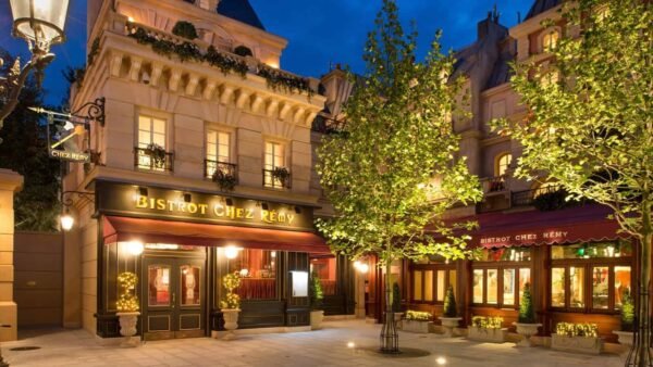 Une charmante vue nocturne du "Bistrot Chez Rémy", l'une des principales attractions de Disneyland Paris, avec des intérieurs chaleureusement éclairés et des lampadaires ornés, entouré d'arbres verts luxuriants dans un