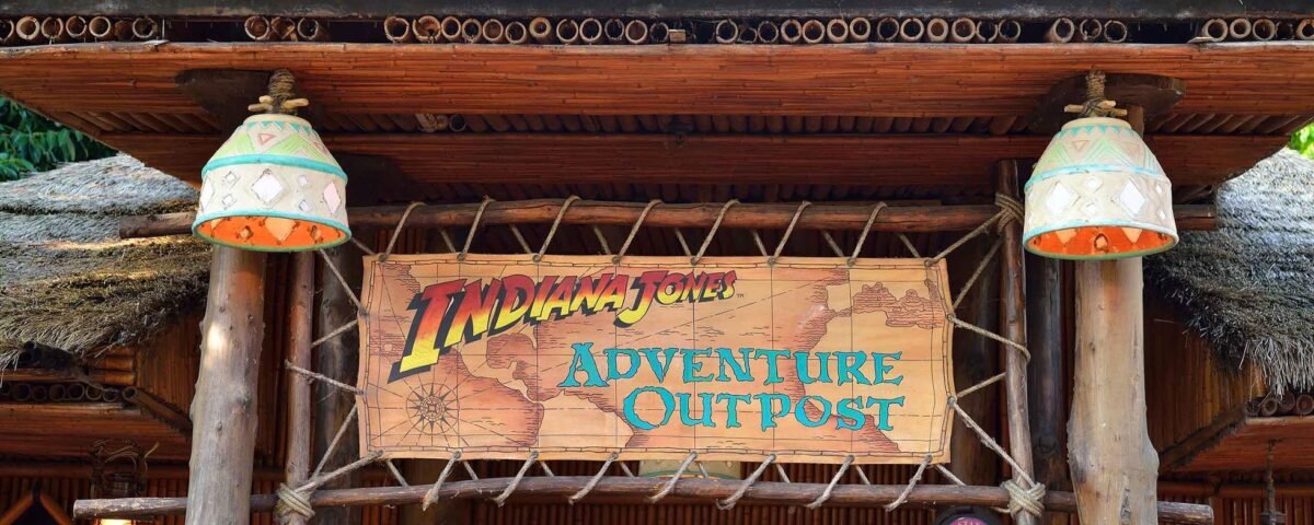 L'entrée de « Indiana Jones Adventure Outpost » à Disneyland Paris présente une pancarte en bois rustique, flanquée de deux lampes suspendues, sous une structure au toit de chaume, avec une verdure luxuriante visible.