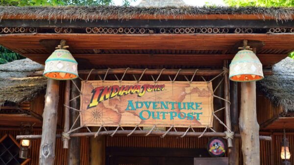 L'entrée de « Indiana Jones Adventure Outpost » à Disneyland Paris présente une pancarte en bois rustique, flanquée de deux lampes suspendues, sous une structure au toit de chaume, avec une verdure luxuriante visible.