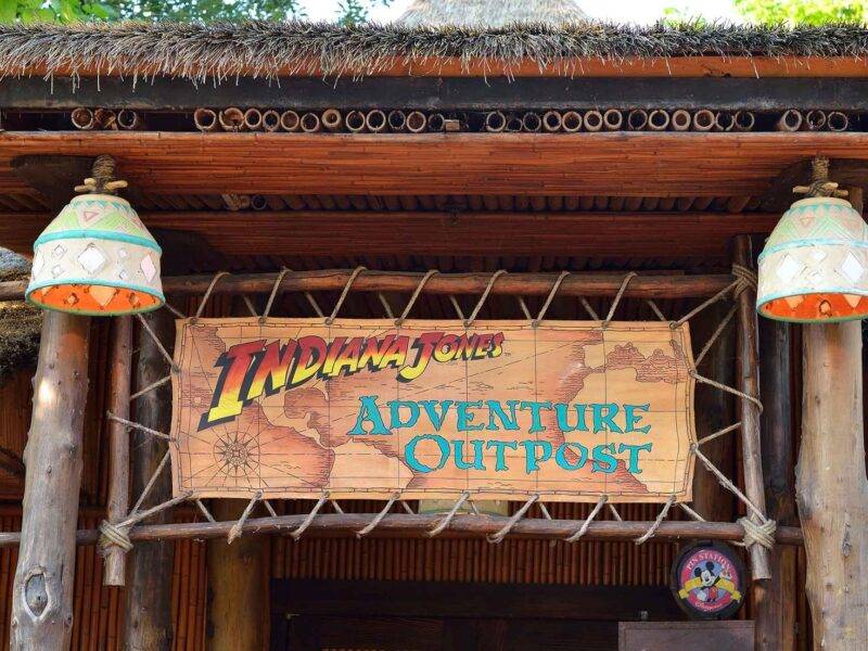 L'entrée de « Indiana Jones Adventure Outpost » à Disneyland Paris présente une pancarte en bois rustique, flanquée de deux lampes suspendues, sous une structure au toit de chaume, avec une verdure luxuriante visible.