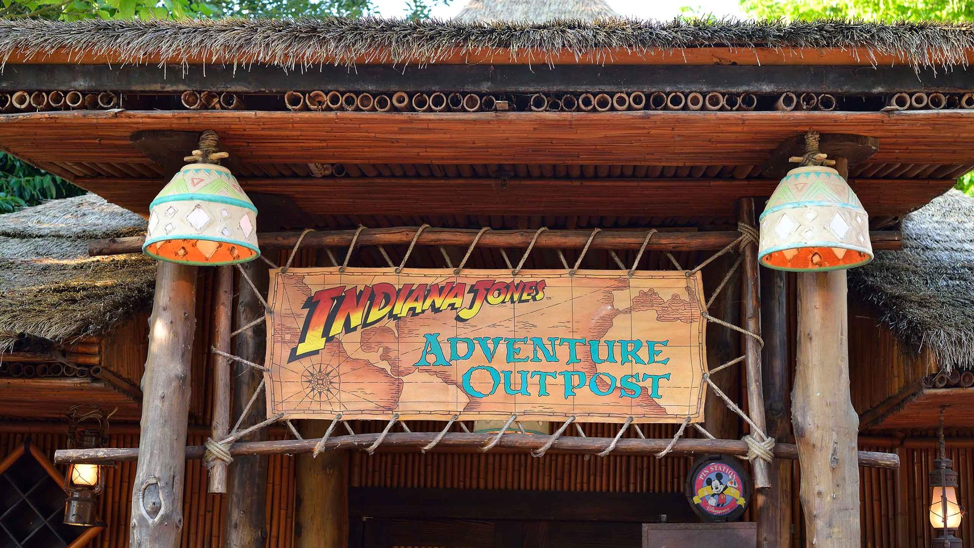 L'entrée de « Indiana Jones Adventure Outpost » à Disneyland Paris présente une pancarte en bois rustique, flanquée de deux lampes suspendues, sous une structure au toit de chaume, avec une verdure luxuriante visible.