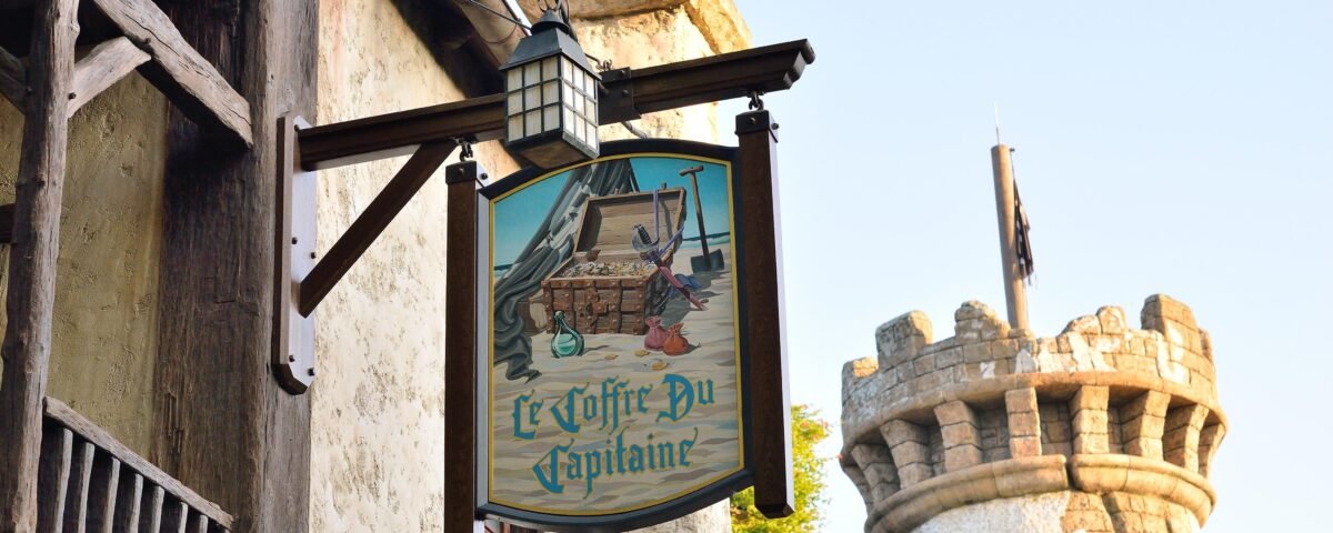 Une enseigne intitulée "Le Coffre du Capitaine" présentant des images nautiques, suspendue à une poutre en bois près d'un bâtiment rustique, avec une tour en pierre vue en arrière-plan.