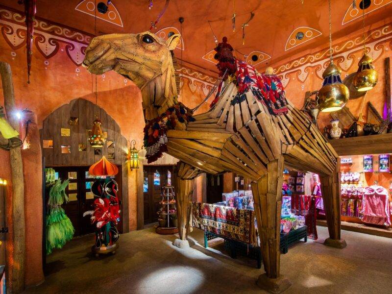 Une boutique vibrante et colorée de Disneyland Paris, Les Trésors de Schéhérazade, présente un modèle de chameau grandeur nature orné de décorations traditionnelles, entouré de lanternes suspendues et d'arcades complexes.