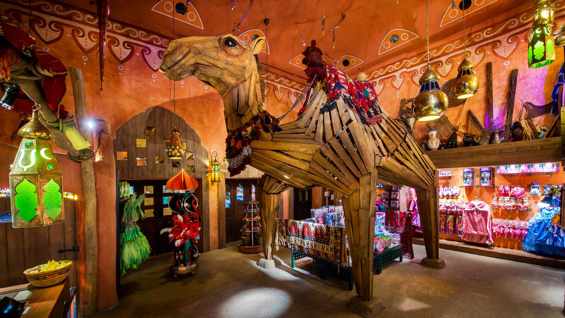 Une boutique vibrante et colorée de Disneyland Paris, Les Trésors de Schéhérazade, présente un modèle de chameau grandeur nature orné de décorations traditionnelles, entouré de lanternes suspendues et d'arcades complexes.