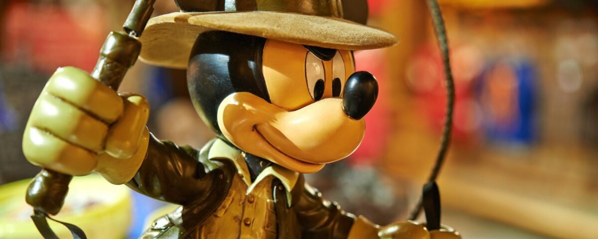 Une figurine détaillée de Mickey Mouse habillée en Indiana Jones, tenant un fouet et portant un fedora, sur un fond coloré flou à Disneyland Paris.