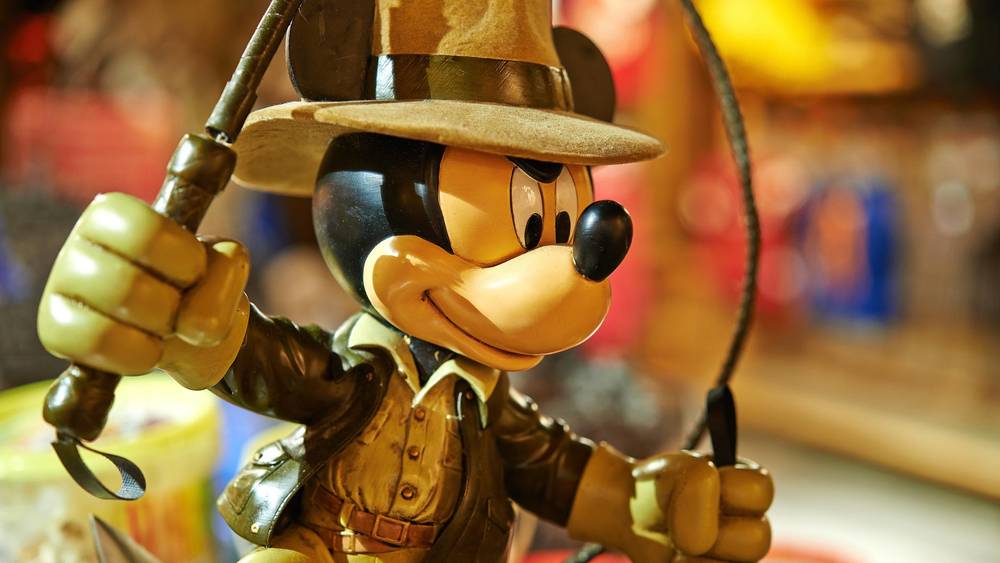 Une figurine détaillée de Mickey Mouse habillée en Indiana Jones, tenant un fouet et portant un fedora, sur un fond coloré flou à Disneyland Paris.