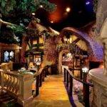Un intérieur fantaisiste représentant une salle au thème fantastique inspirée de Disneyland Paris, avec des structures en forme d'arbre, des toits couverts de mousse et une allée pavée menant à un bar aux allures de château avec des vitraux