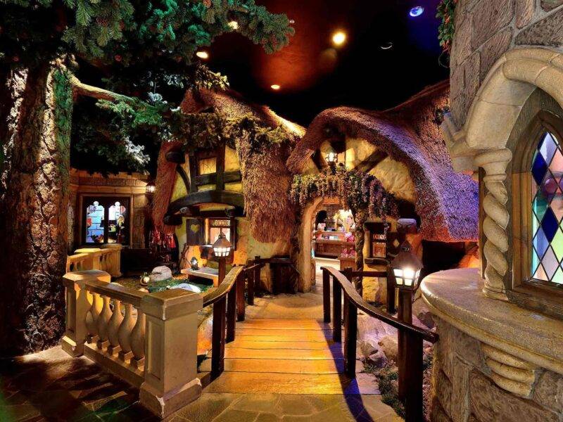 Un intérieur fantaisiste représentant une salle au thème fantastique inspirée de Disneyland Paris, avec des structures en forme d'arbre, des toits couverts de mousse et une allée pavée menant à un bar aux allures de château avec des vitraux