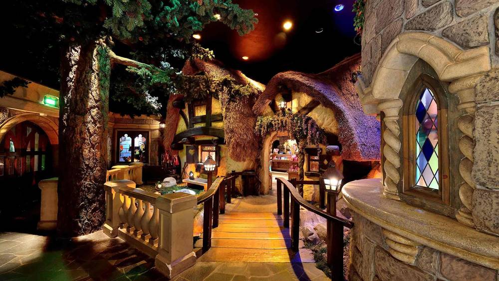 Un intérieur fantaisiste représentant une salle au thème fantastique inspirée de Disneyland Paris, avec des structures en forme d'arbre, des toits couverts de mousse et une allée pavée menant à un bar aux allures de château avec des vitraux