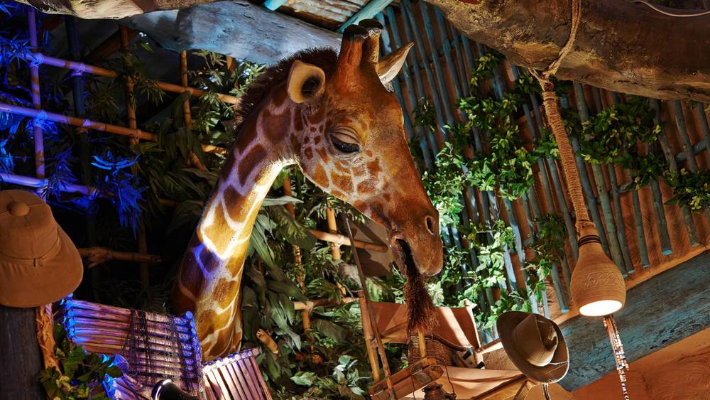 Une girafe se nourrissant de plantes dans une salle au thème rustique de Disneyland Paris avec un éclairage chaleureux et diverses décorations suspendues comme des chapeaux et des paniers.