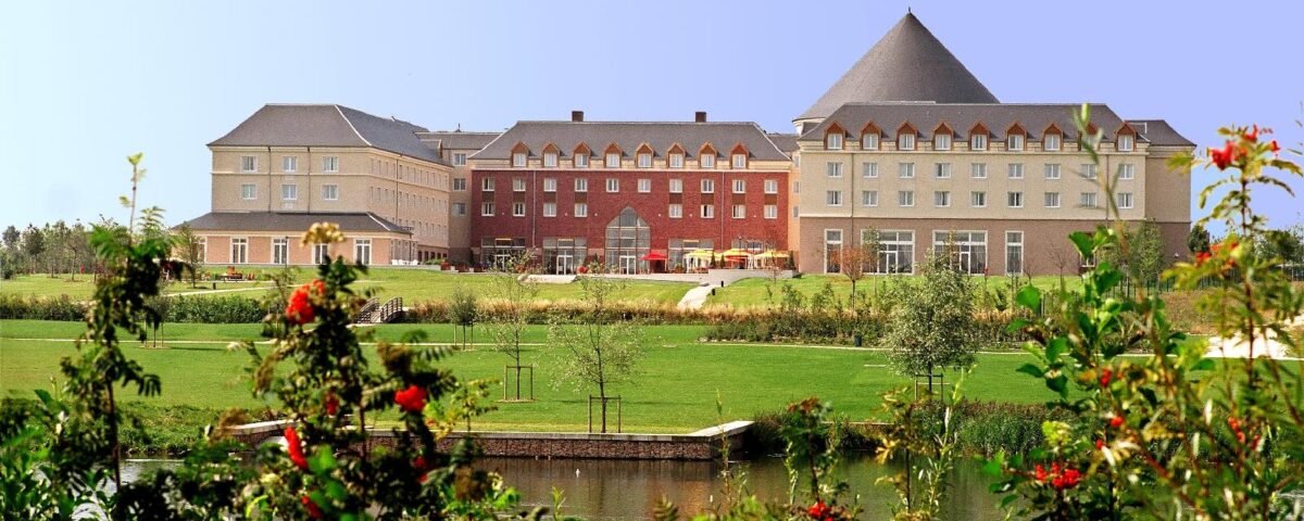 Grand et élégant Grand Magic Hotel avec des lignes de toit variées surplombant un étang tranquille, entouré de pelouses verdoyantes et de fleurs éclatantes au premier plan.