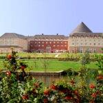 Grand et élégant Grand Magic Hotel avec des lignes de toit variées surplombant un étang tranquille, entouré de pelouses verdoyantes et de fleurs éclatantes au premier plan.