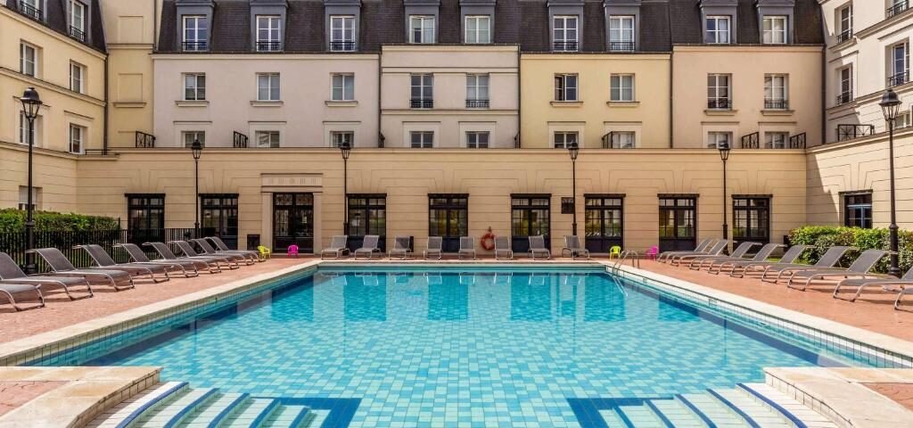 Une cour d'hôtel luxueuse dotée d'une piscine d'un bleu étincelant entourée de nombreuses chaises longues, avec l'élégant bâtiment hôtelier à plusieurs étages Hipark by Adagio en arrière-plan sous un ciel dégagé.