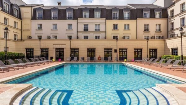 Une cour d'hôtel luxueuse dotée d'une piscine d'un bleu étincelant entourée de nombreuses chaises longues, avec l'élégant bâtiment hôtelier à plusieurs étages Hipark by Adagio en arrière-plan sous un ciel dégagé.