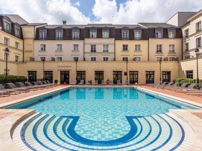 Une cour d'hôtel luxueuse dotée d'une piscine d'un bleu étincelant entourée de nombreuses chaises longues, avec l'élégant bâtiment hôtelier à plusieurs étages Hipark by Adagio en arrière-plan sous un ciel dégagé.