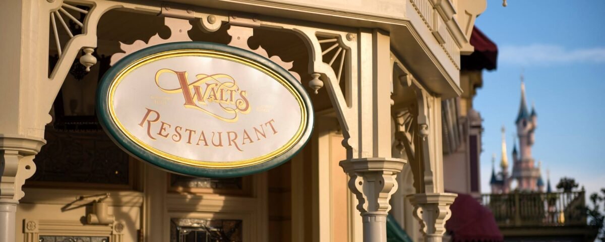 Enseigne élégante pour "Walt's Restaurant" avec une écriture ornée sur une plaque ovale, montée sur une façade classique de couleur crème à Disneyland Paris. Un château de conte de fées lointain est visible dans