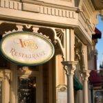 Enseigne élégante pour "Walt's Restaurant" avec une écriture ornée sur une plaque ovale, montée sur une façade classique de couleur crème à Disneyland Paris. Un château de conte de fées lointain est visible dans