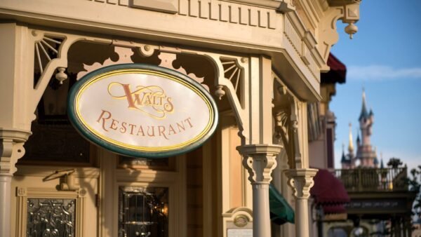 Enseigne élégante pour "Walt's Restaurant" avec une écriture ornée sur une plaque ovale, montée sur une façade classique de couleur crème à Disneyland Paris. Un château de conte de fées lointain est visible dans