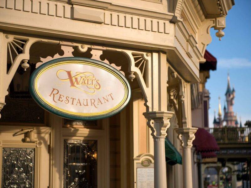 Enseigne élégante pour "Walt's Restaurant" avec une écriture ornée sur une plaque ovale, montée sur une façade classique de couleur crème à Disneyland Paris. Un château de conte de fées lointain est visible dans