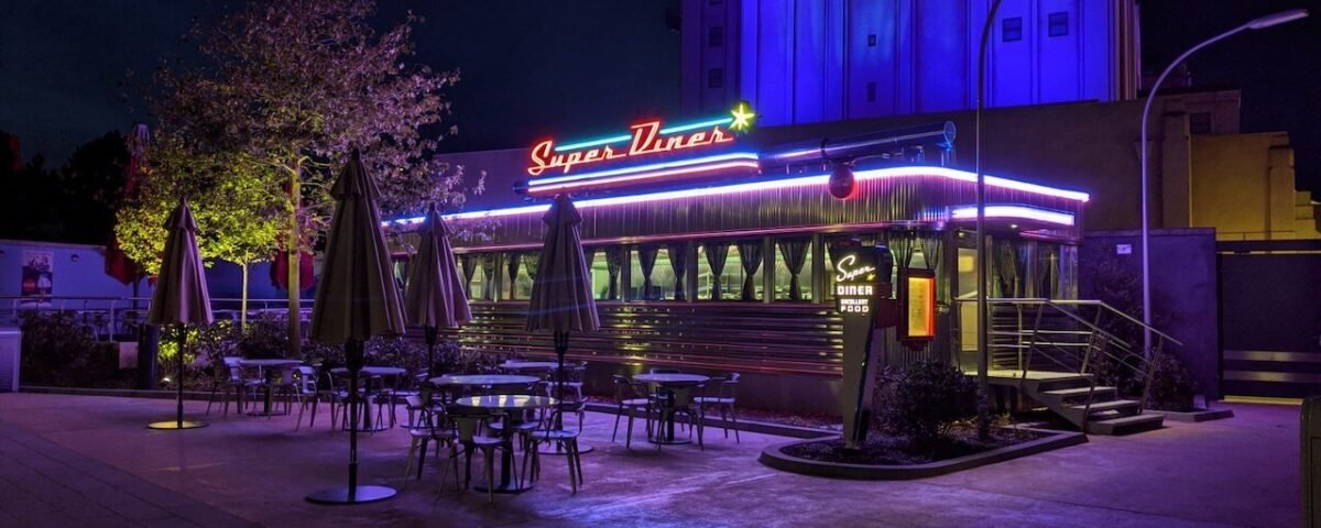 Un restaurant de style rétro nommé "super diner", éclairé la nuit par des néons bleus et rouges, doté de sièges extérieurs sous des parasols, dans une ambiance faiblement éclairée.