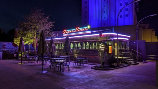 Un restaurant de style rétro nommé "super diner", éclairé la nuit par des néons bleus et rouges, doté de sièges extérieurs sous des parasols, dans une ambiance faiblement éclairée.