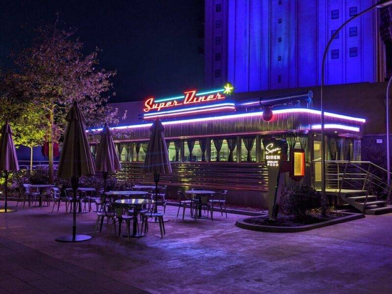 Un restaurant de style rétro nommé "super diner", éclairé la nuit par des néons bleus et rouges, doté de sièges extérieurs sous des parasols, dans une ambiance faiblement éclairée.
