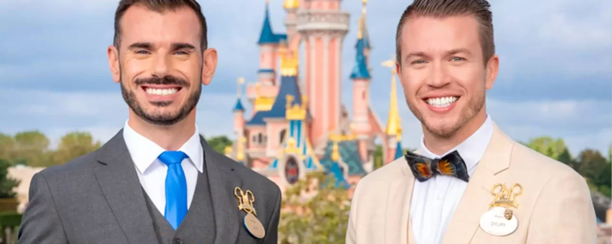 Deux ambassadeurs souriants portant des badges Disneyland Paris, l'un en costume gris et cravate bleue, l'autre en costume beige avec un nœud papillon coloré, debout devant un Disney