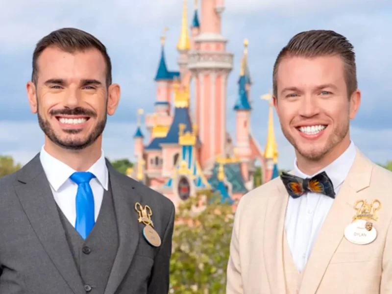 Deux ambassadeurs souriants portant des badges Disneyland Paris, l'un en costume gris et cravate bleue, l'autre en costume beige avec un nœud papillon coloré, debout devant un Disney