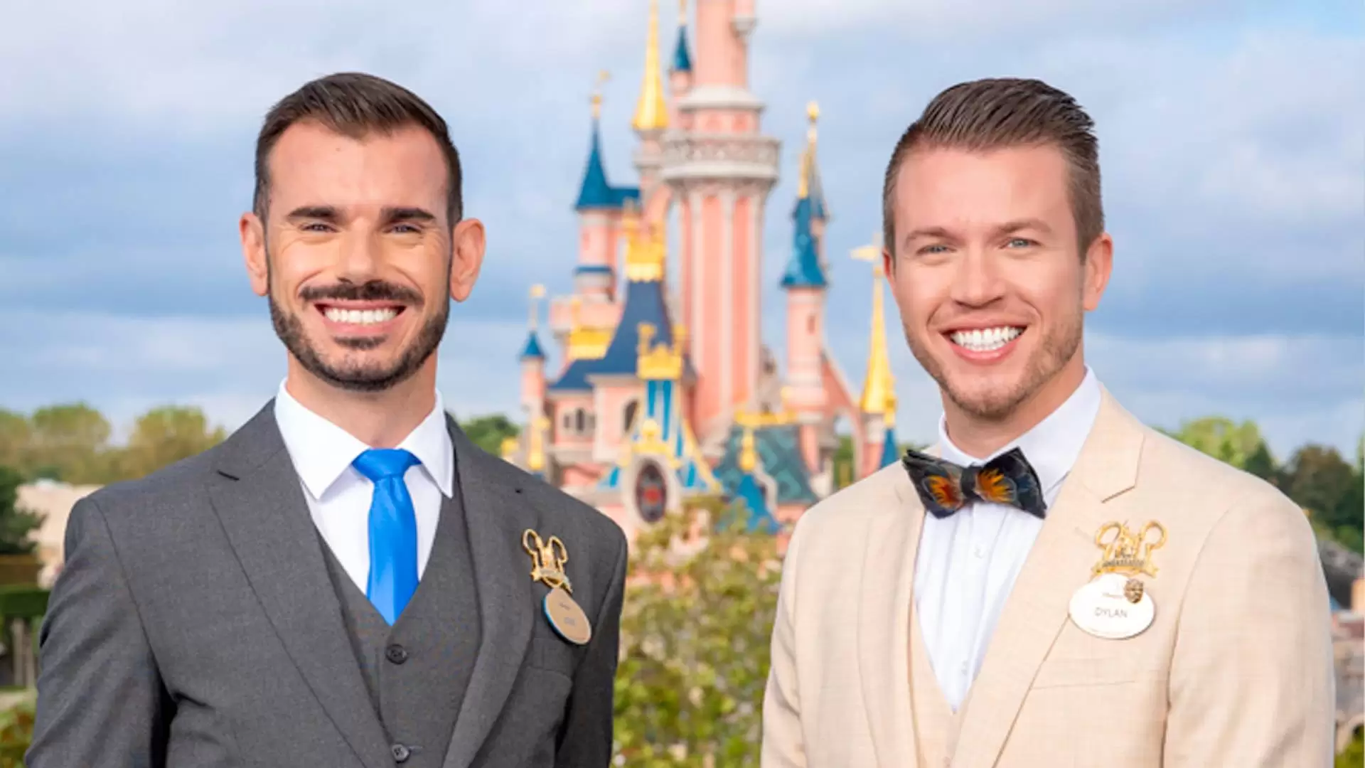Deux ambassadeurs souriants portant des badges Disneyland Paris, l'un en costume gris et cravate bleue, l'autre en costume beige avec un nœud papillon coloré, debout devant un Disney