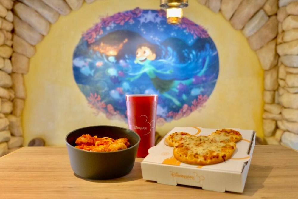 Une pizza et une boisson de la Pizzeria Bella Notte sur une table à côté d'une fresque murale à Disneyland Paris.