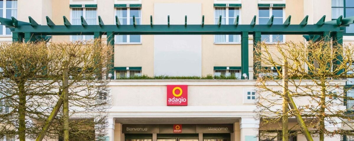 Vue de face de l'entrée de l'hôtel Disneyland Paris Adagio présentant une façade symétrique avec de grandes fenêtres, décorée de treillis verts et de haies soigneusement taillées, et un panneau de bienvenue en plusieurs langues.