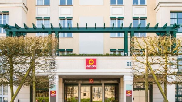 Vue de face de l'entrée de l'hôtel Disneyland Paris Adagio présentant une façade symétrique avec de grandes fenêtres, décorée de treillis verts et de haies soigneusement taillées, et un panneau de bienvenue en plusieurs langues.