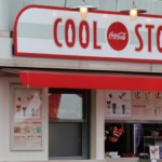 Un kiosque de restauration rapide classique appelé « cool stop » situé dans Disney Village avec un logo Coca-Cola, avec une palette de couleurs rouge et blanche, des panneaux de menu visibles et une glacière à boissons à l'intérieur.