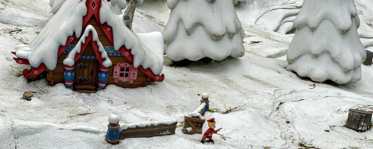 Une scène hivernale fantaisiste mettant en vedette un cottage coloré et décoré avec un toit enneigé et des conifères enneigés, un modèle d'enfant en luge à proximité, le tout dans un paysage enneigé miniature rappelant Disneyland Paris.