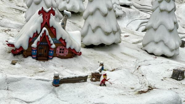 Une scène hivernale fantaisiste mettant en vedette un cottage coloré et décoré avec un toit enneigé et des conifères enneigés, un modèle d'enfant en luge à proximité, le tout dans un paysage enneigé miniature rappelant Disneyland Paris.