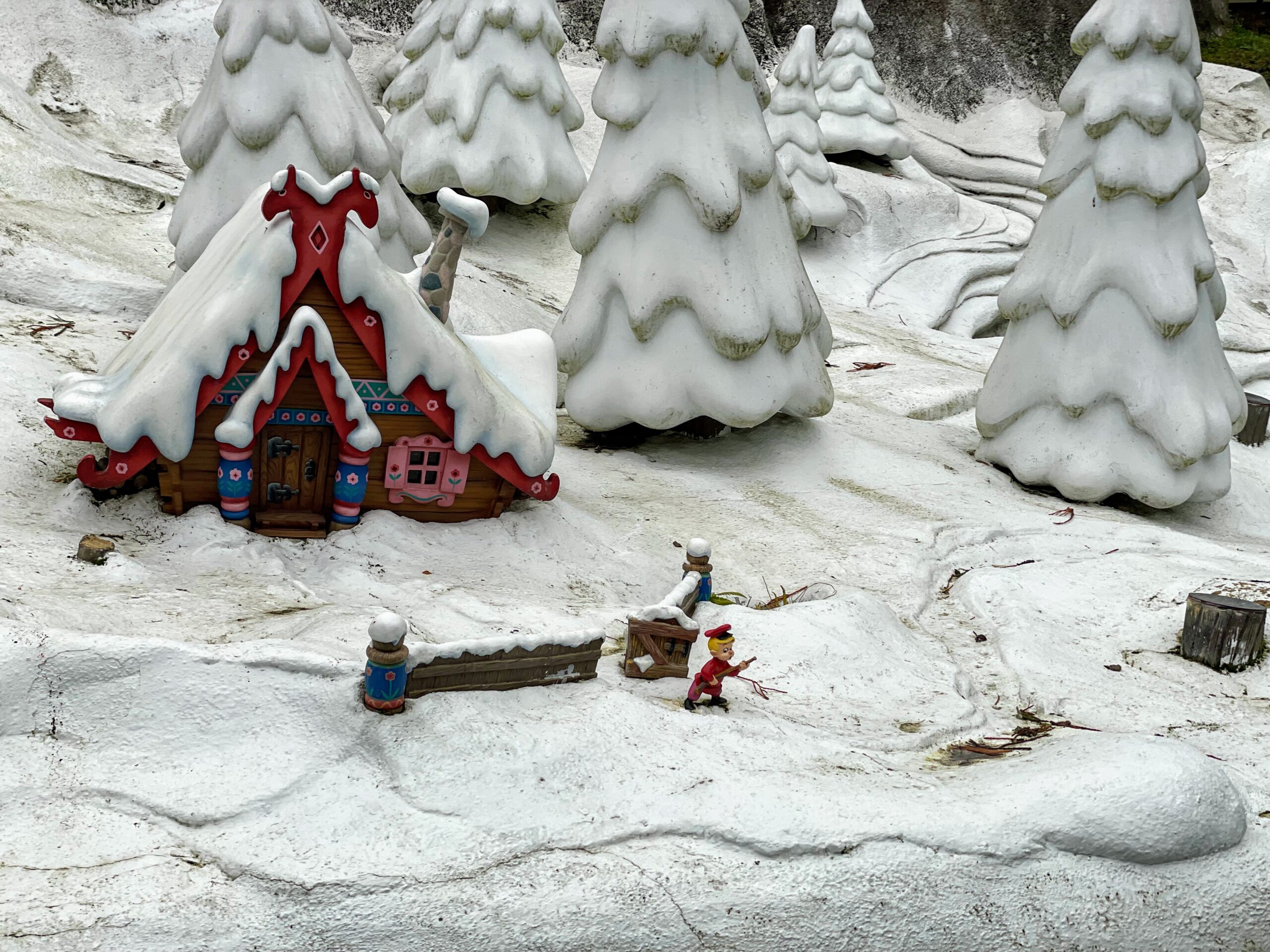 Une scène hivernale fantaisiste mettant en vedette un cottage coloré et décoré avec un toit enneigé et des conifères enneigés, un modèle d'enfant en luge à proximité, le tout dans un paysage enneigé miniature rappelant Disneyland Paris.