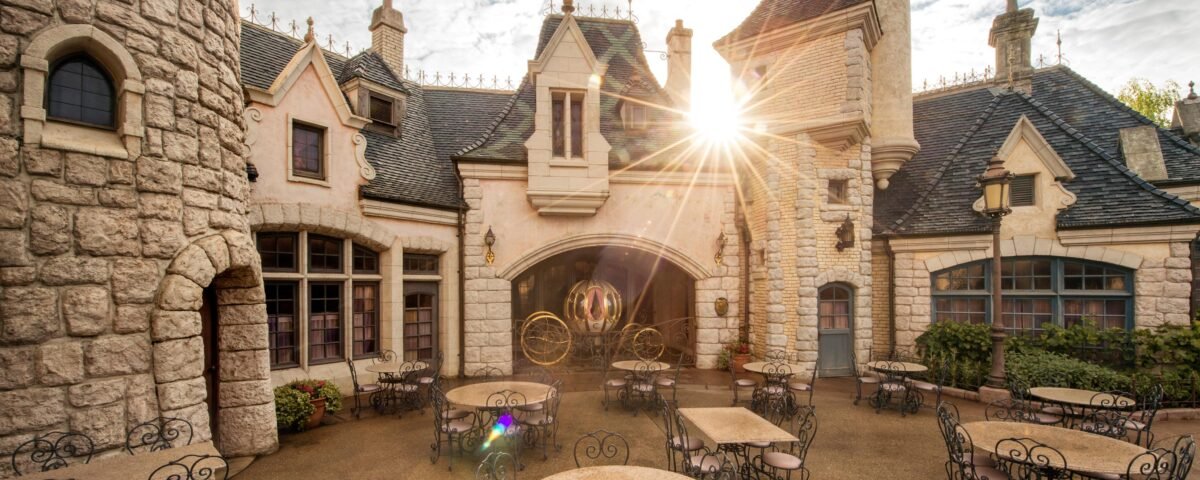 Lever de soleil sur la charmante Auberge de Cendrillon à Disneyland Paris, avec ses murs en pierre et ses détails architecturaux complexes, avec ses sièges extérieurs et ses rayons de soleil traversant une arcade.