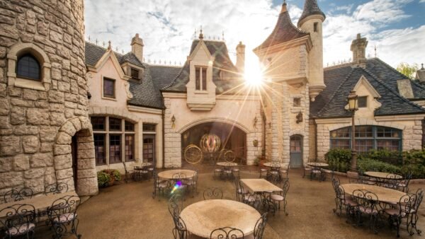 Lever de soleil sur la charmante Auberge de Cendrillon à Disneyland Paris, avec ses murs en pierre et ses détails architecturaux complexes, avec ses sièges extérieurs et ses rayons de soleil traversant une arcade.