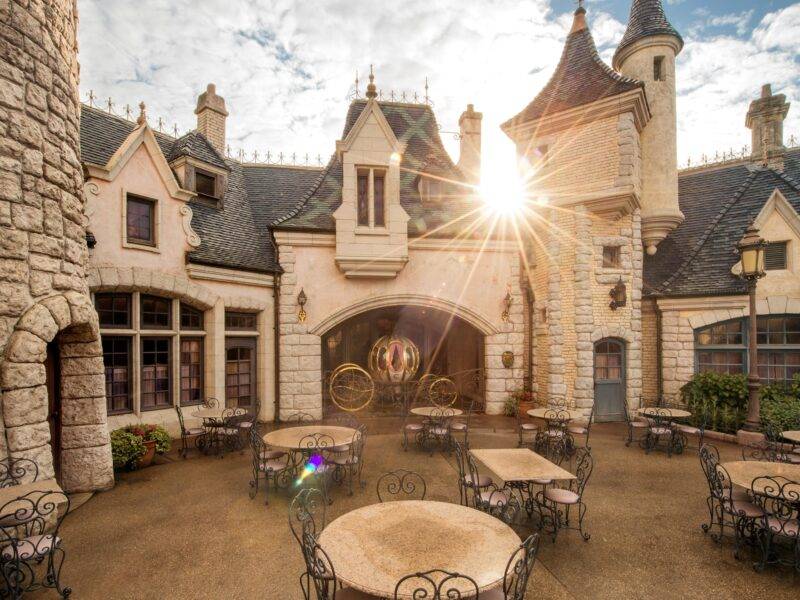 Lever de soleil sur la charmante Auberge de Cendrillon à Disneyland Paris, avec ses murs en pierre et ses détails architecturaux complexes, avec ses sièges extérieurs et ses rayons de soleil traversant une arcade.