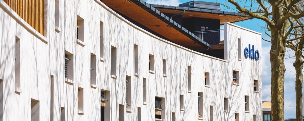 Bâtiment blanc moderne avec des motifs de façade abstraits et le logo « eklo », accompagné d'un arbre nu et d'un ciel bleu.