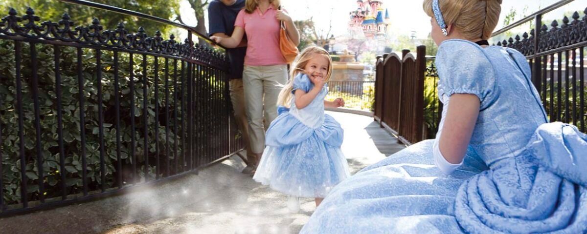 Une jeune fille en costume de princesse court avec enthousiasme vers une femme habillée en Cendrillon en robe bleue à l'Auberge de Cendrillon, avec sa famille souriant derrière elle sous le soleil